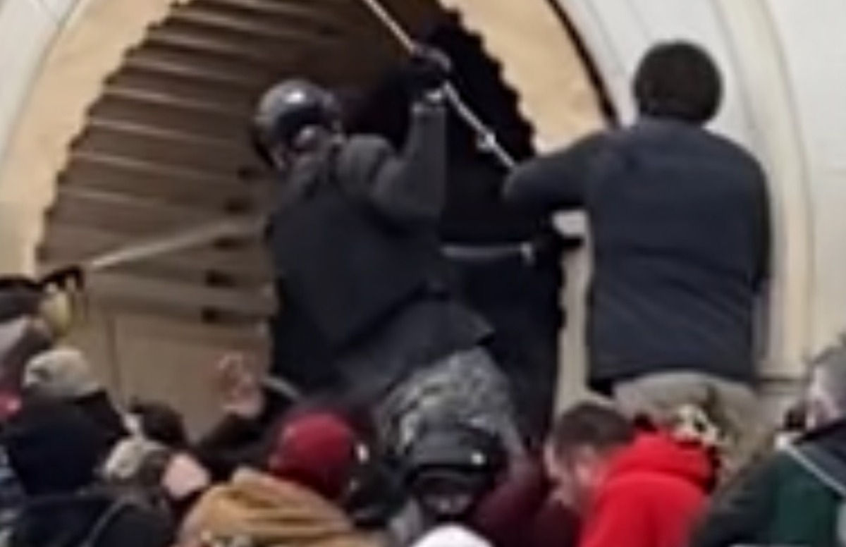 A man wearing a similar black helmet and face covering provides base support and what appears to be protection for the individual with the pole to attack the door area – Source: National File